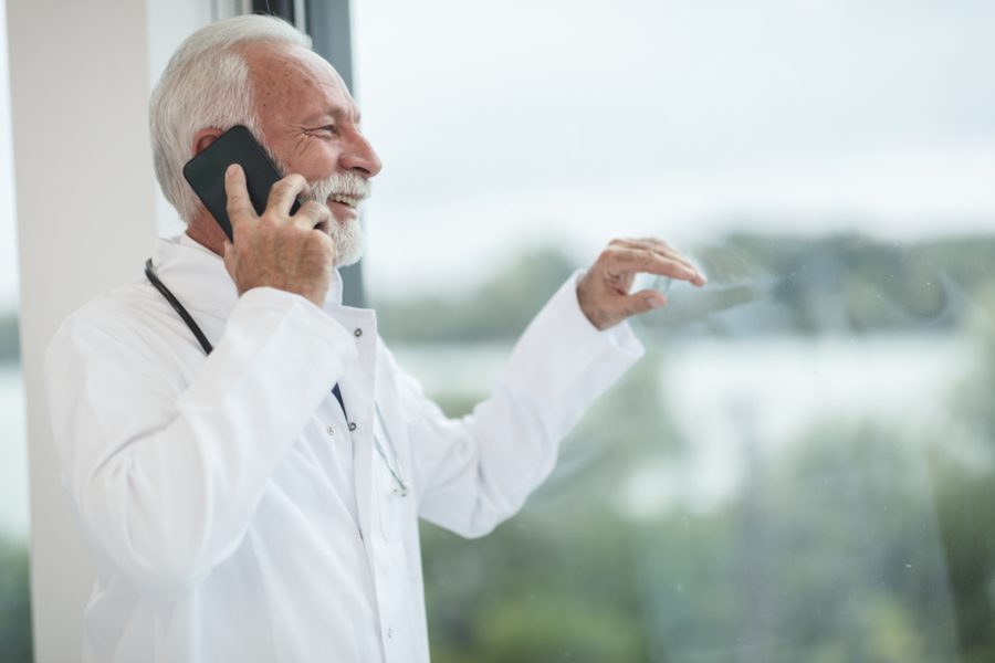 A grey-haired man in a white coat with a stethoscope laughing on the phone by a window