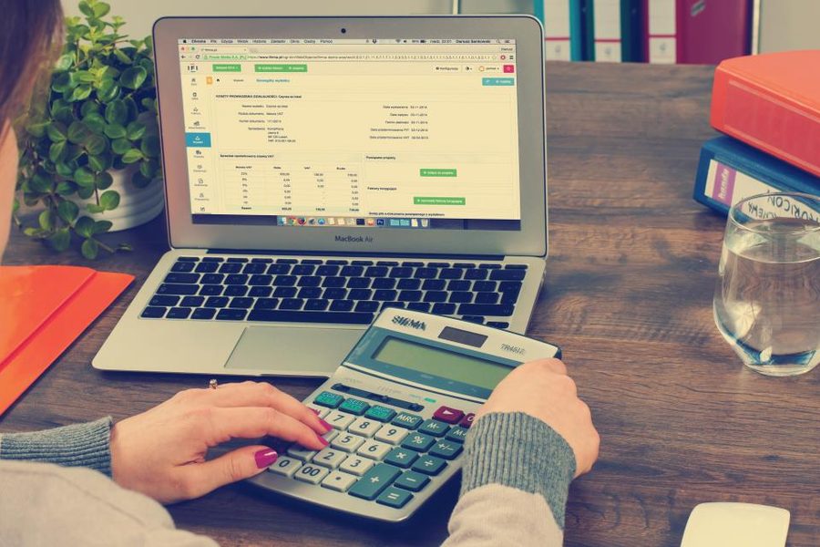 A woman’s hands on a calculator and laptop keyboard at a wooden desk with folders and a plant