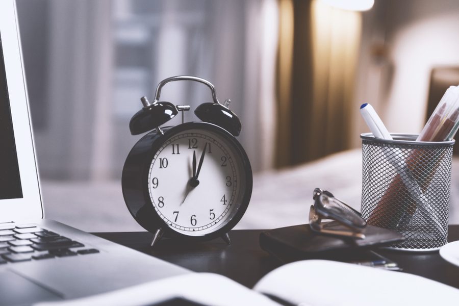 A black alarm clock on a desk beside a laptop and a cup of pens