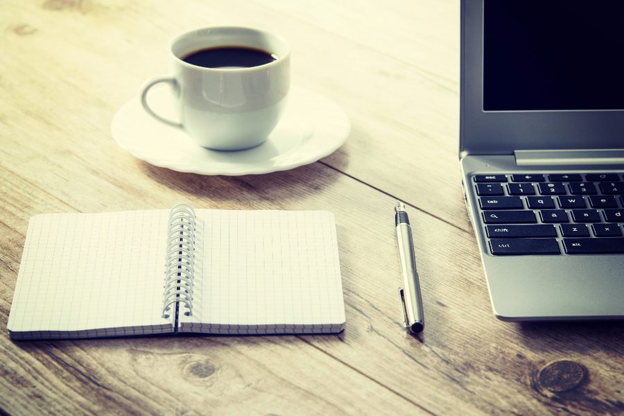 A cup of coffee, an open notebook and pen beside a laptop on a wooden desk