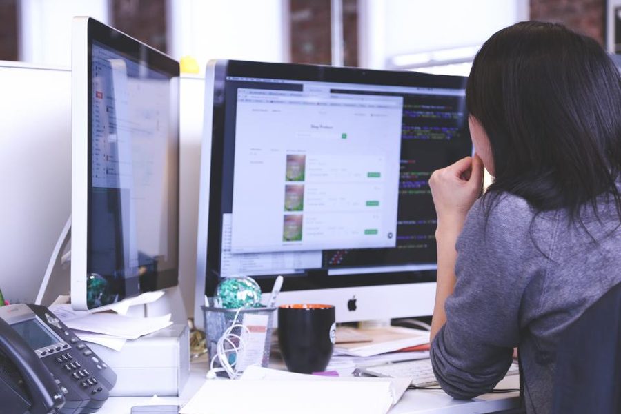 A woman at a desk looking at two computer monitors showing a task list