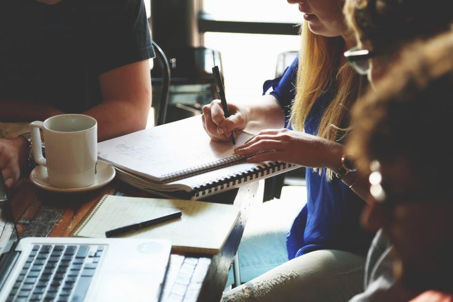 Colleagues around a table with coffee cups and a notebook, one writing while others talk