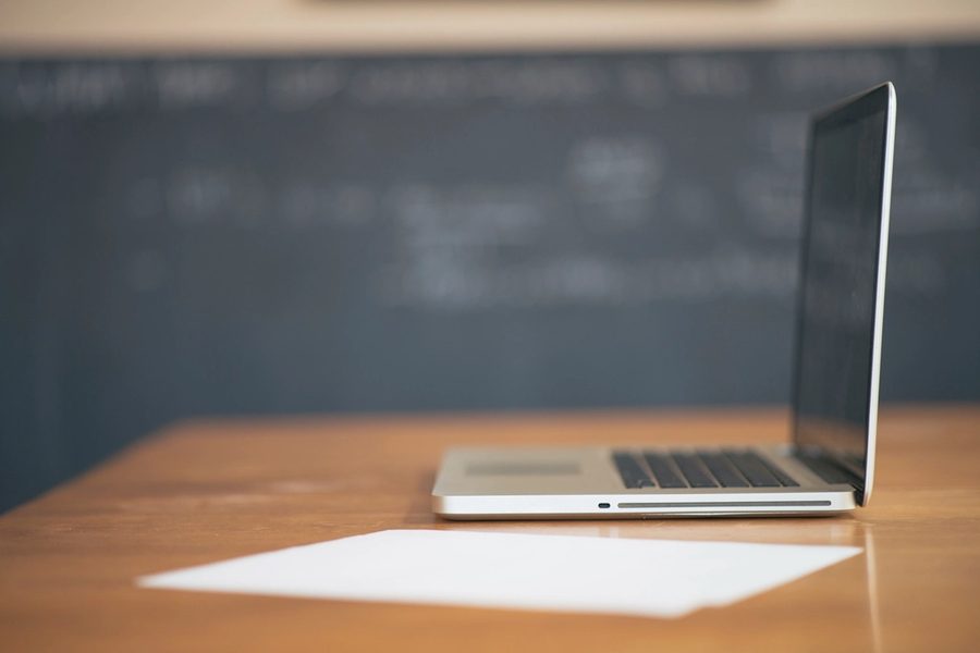 A laptop open on a wooden desk beside a blank sheet of paper, a chalkboard blurred behind it