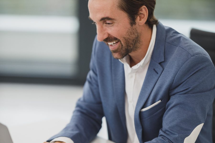 A smiling man in a blue blazer sitting at a desk near a window