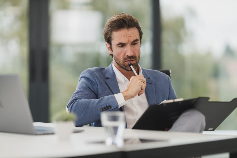 A man in a blazer holding a pen to his chin while looking at papers on a clipboard at a desk