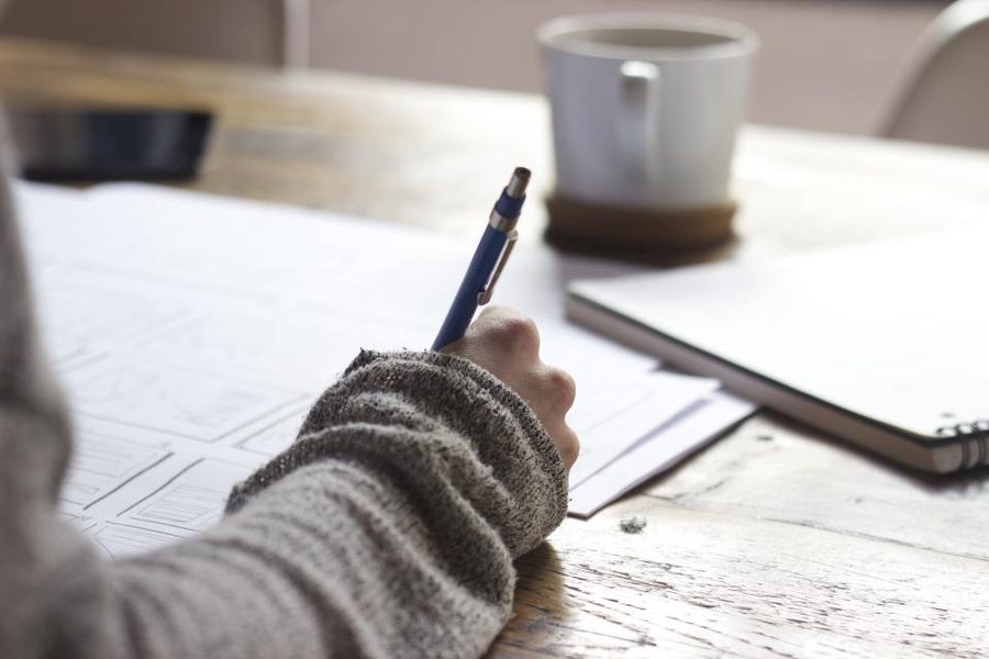 A hand writing with a pen on paper beside a cup of coffee on a wooden desk