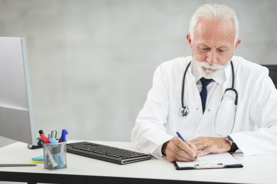A grey-haired man in a white coat and stethoscope writing at a desk