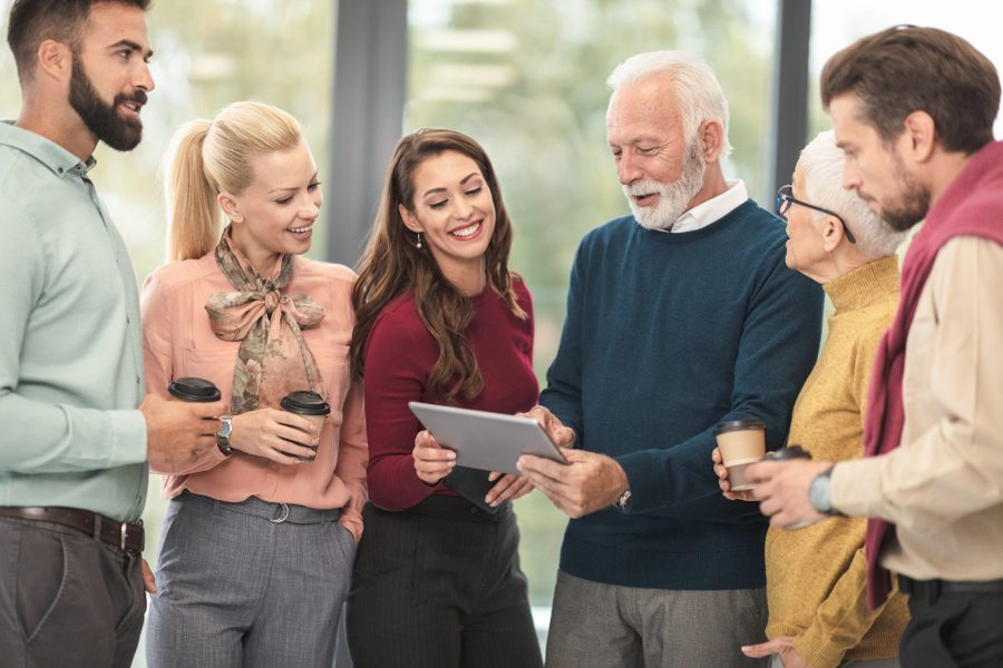 Six colleagues of different ages standing together looking at a tablet one of them is holding