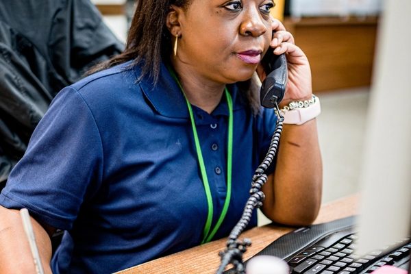 A woman in a polo shirt and lanyard answering a desk phone beside a computer monitor